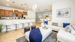 A modern living room with a sofa, coffee table, and bar stools. There is a kitchen area with wooden cabinets and a microwave.