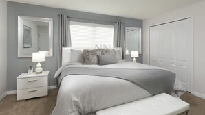 A bedroom with a bed, a nightstand, a bench, a window with curtains, a mirror, and a door.