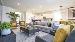 A modern living room with a grey sofa, coffee table, and a dining area with a kitchen in the background. - Last Minute Disney! Private Pool, Hot Tub, Game Room & Themed Bedrooms. Book Now! #107763 (Orlando)