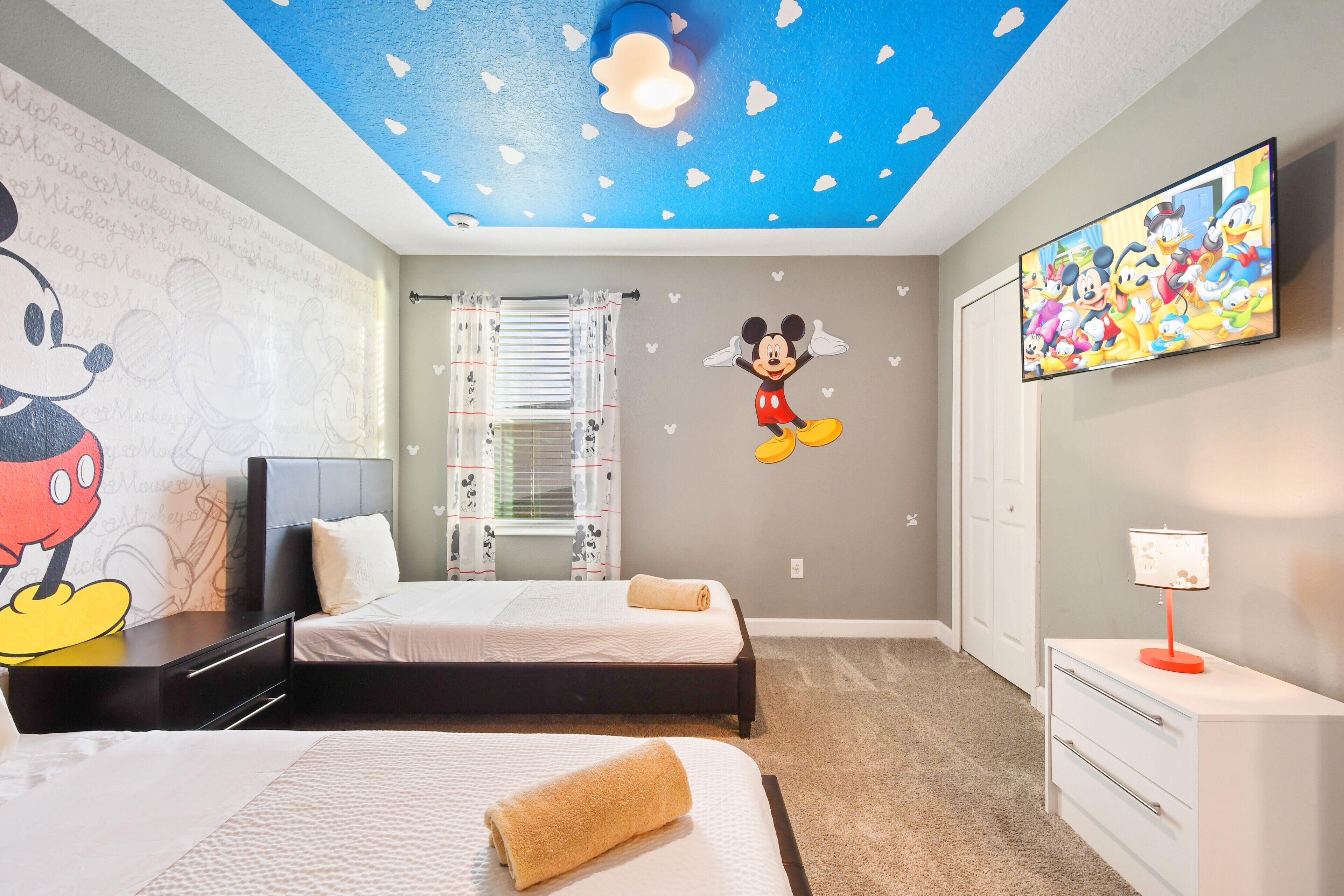 Une chambre d’hôtel avec deux lits, un plafond décoré de motifs étoilés, des autocollants muraux représentant Mickey Mouse et Donald Duck, une télévision et un bureau avec une lampe.