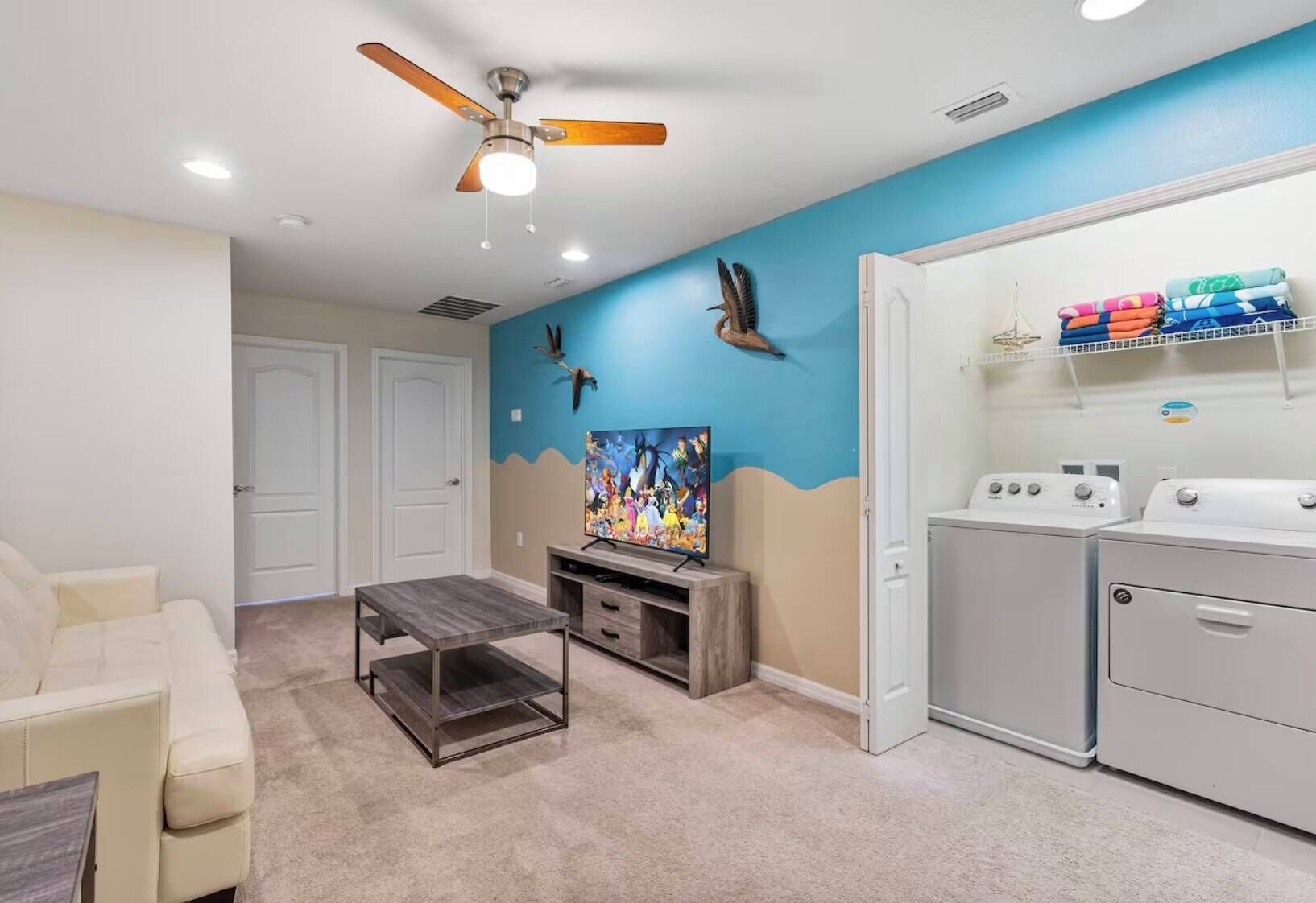 A living room with a TV, a ceiling fan, a coffee table, a sofa, and a laundry area with washing machines.