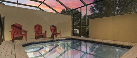 An indoor swimming pool with a glass ceiling, three Adirondack chairs, and a view of trees through the glass.