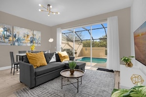 Uma sala de estar moderna com um sofá cinzento, uma mesa de centro redonda e uma grande janela com vista para uma piscina.