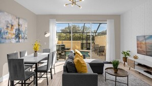 A modern living room with a dining area, a sofa with cushions, a TV, and a large window overlooking a pool and outdoor seating. - Last Minute Disney Getaway! Townhouse with Private Pool. Book Now! #66057 (Orlando)