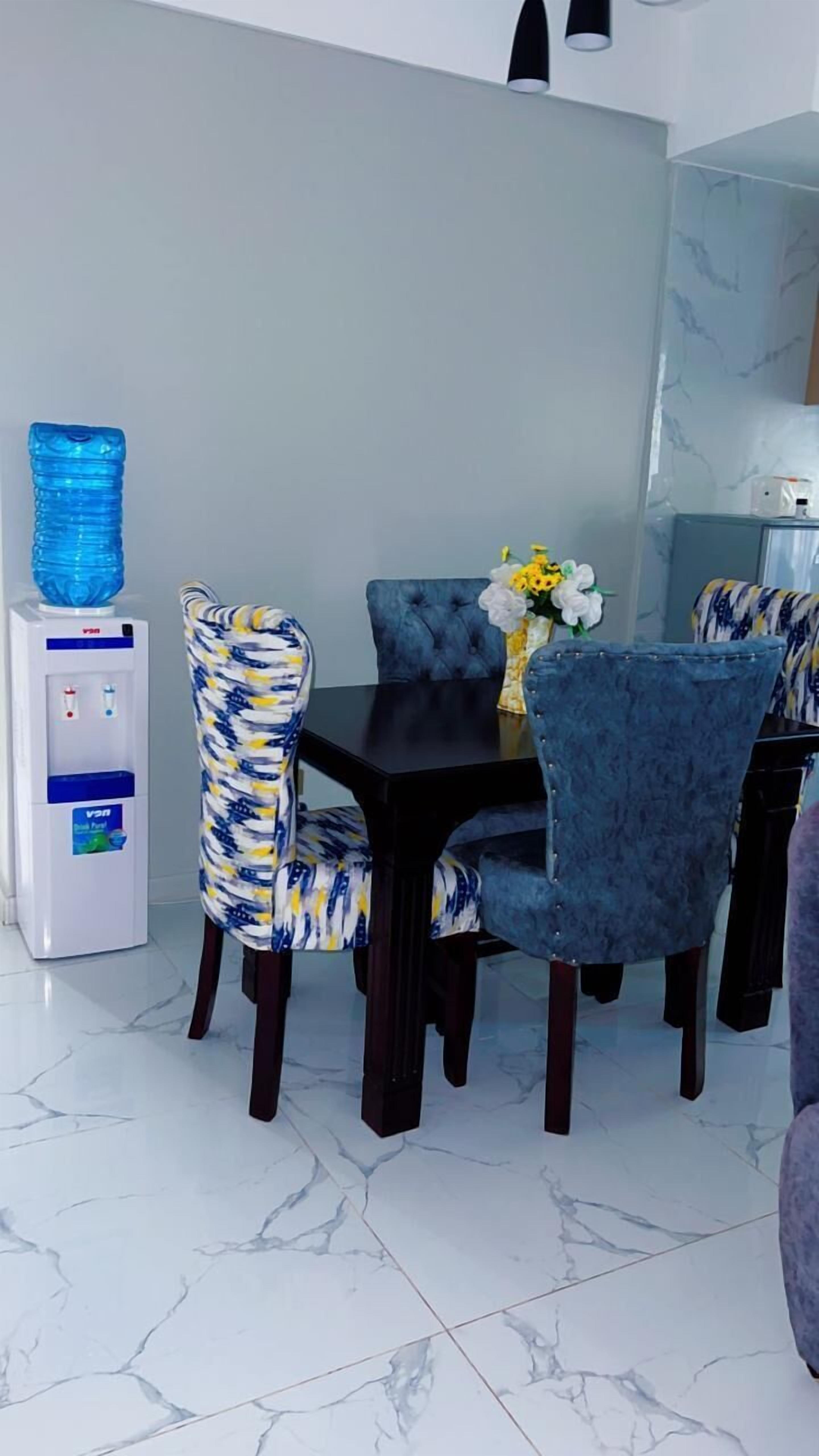 Un coin repas avec une table noire, des chaises recouvertes de tissu bleu et un distributeur d’eau.