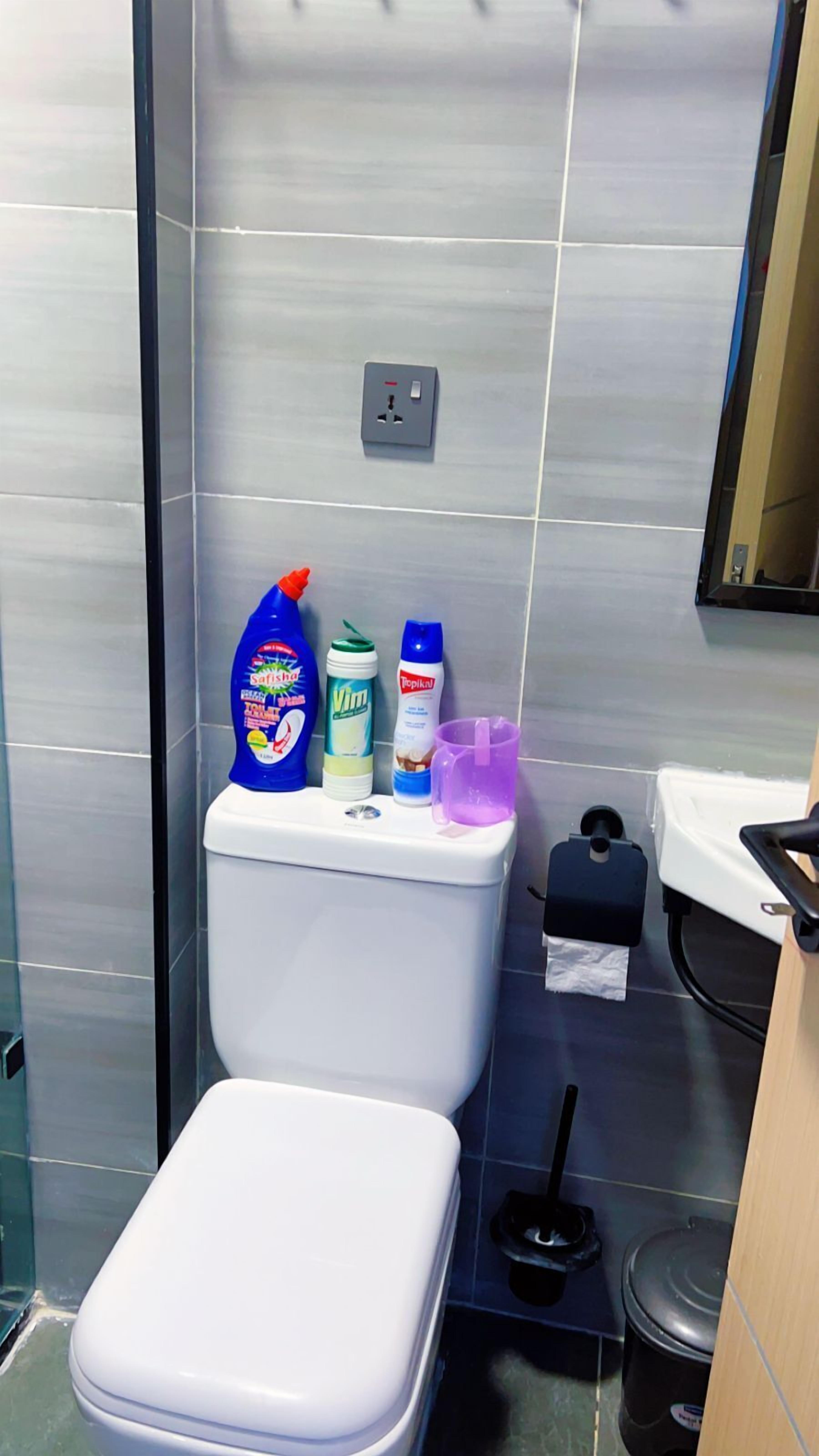 A bathroom with a toilet, a trash bin, and a shelf with cleaning supplies.