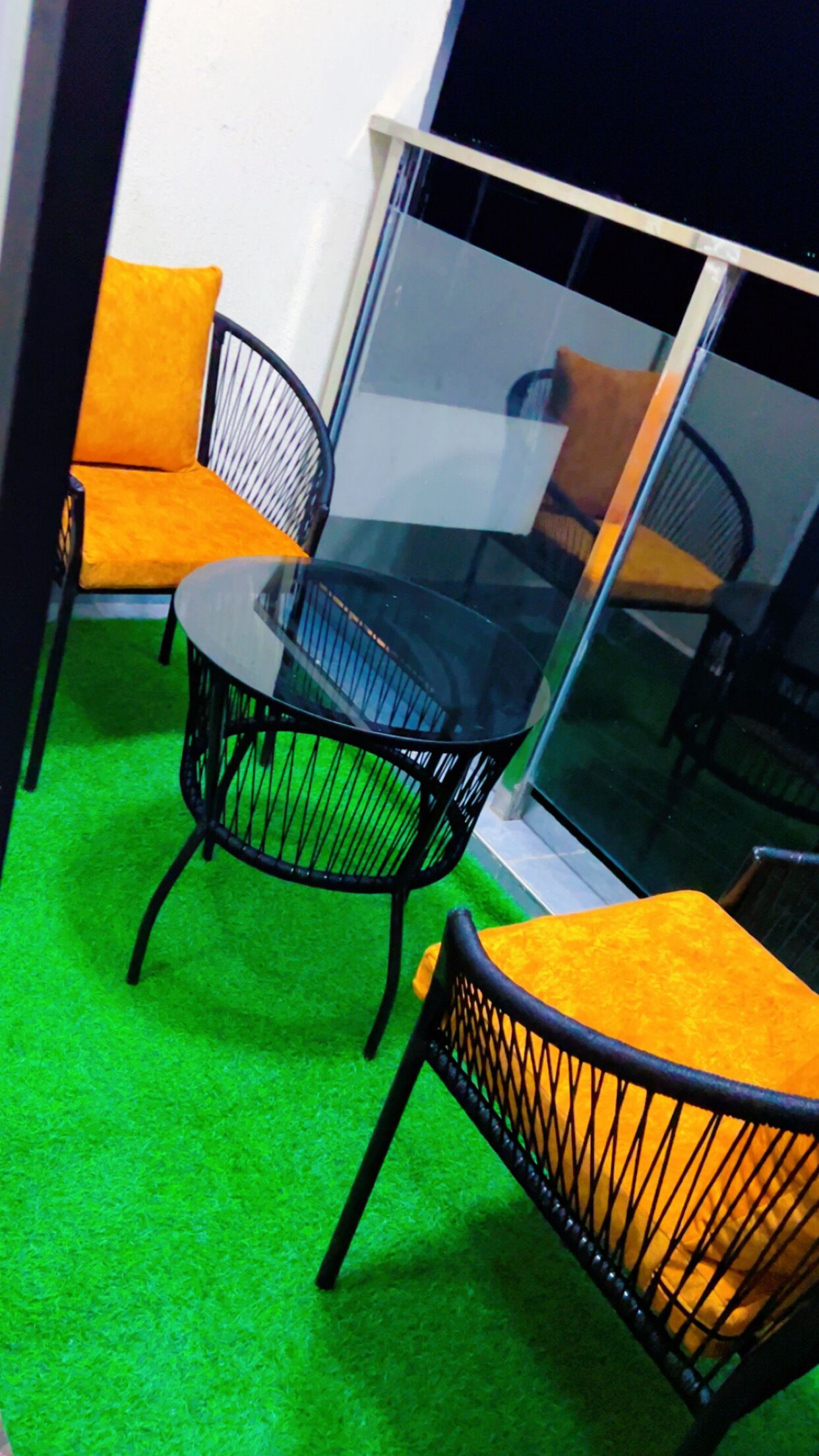 Una habitación con una mesa de vidrio, dos sillas con cojines amarillos, una alfombra verde y un ventanal grande.