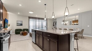 A modern kitchen with dark cabinetry, a large island, pendant lighting, and a dining area with chairs.