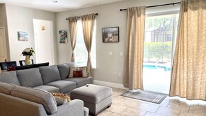 Una sala de estar con un sofá modular, un reposapiés y una mesa de comedor con sillas. Hay dos ventanas con cortinas, una puerta y vista a una piscina.