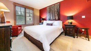 A bedroom with a large bed, a red accent wall, and a window with blinds. - Catamount Lodge 305A - Village Junior Studio (Truckee)