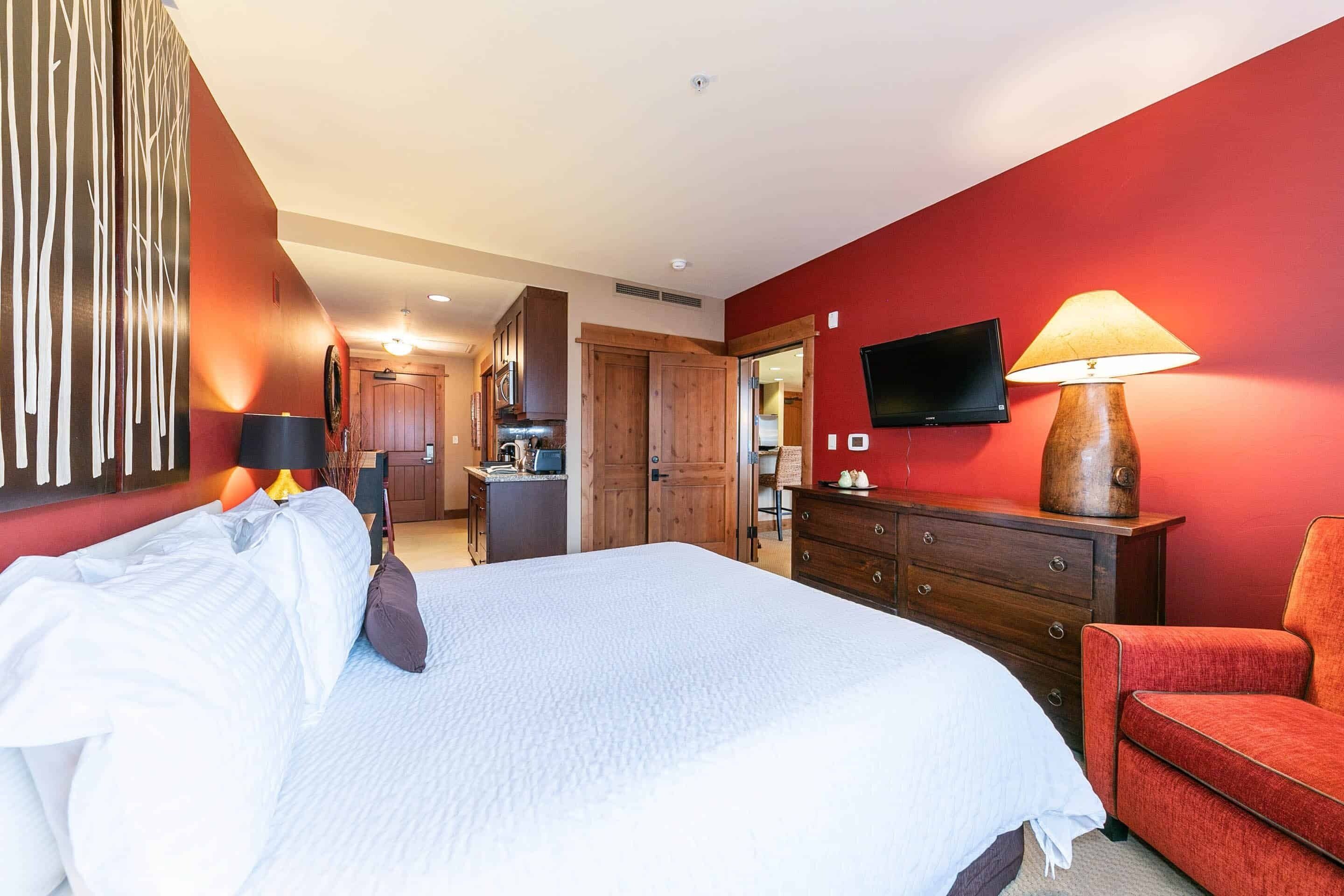 A hotel room with a large bed, a red wall, a wooden dresser, a red armchair, a TV mounted on the wall, and a kitchen area in the background.