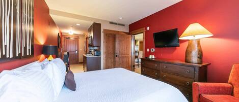 A hotel room with a large bed, a red wall, a wooden dresser, a red armchair, a TV mounted on the wall, and a kitchen area in the background.