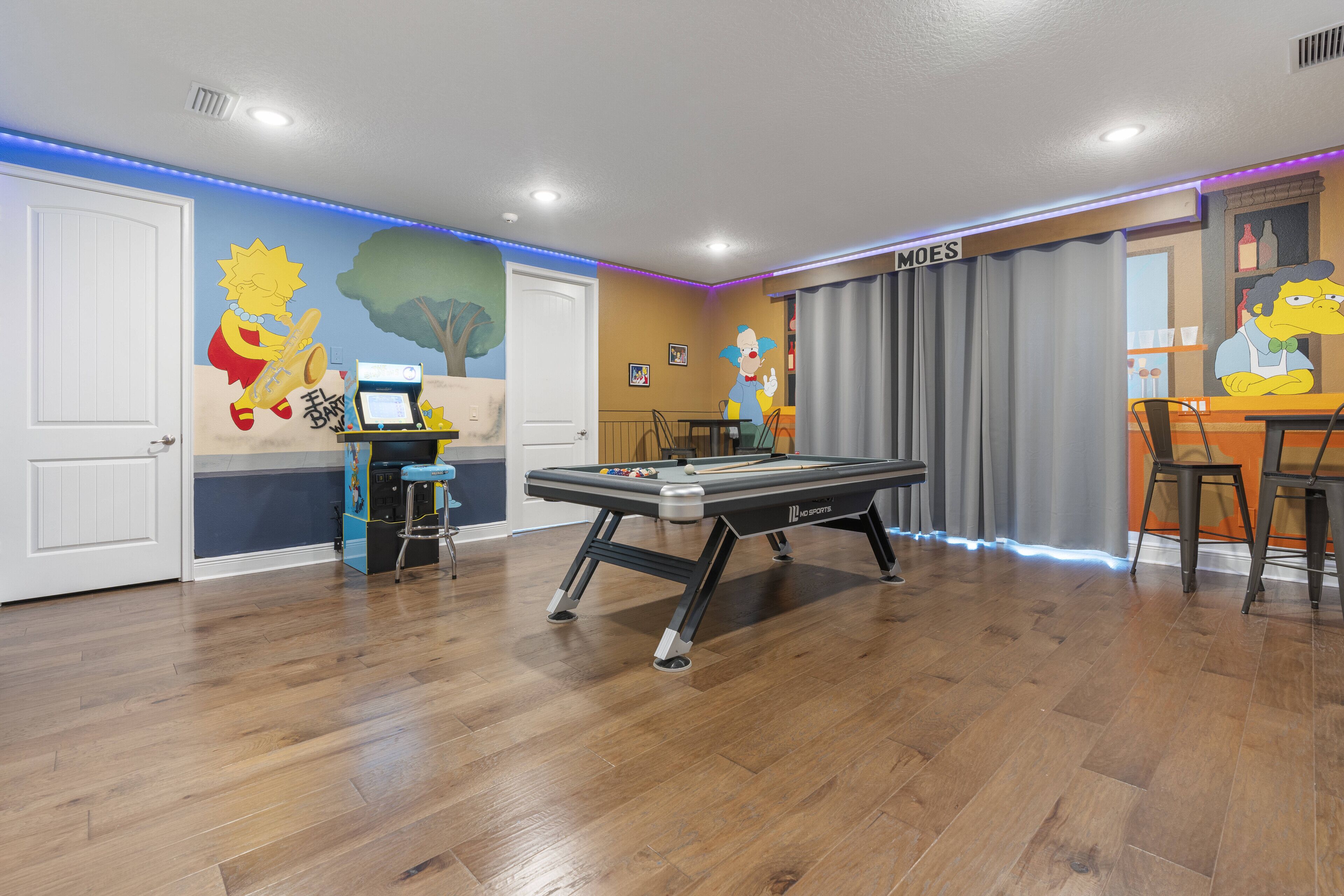 Una mesa de billar en una habitación con suelo de madera, una zona de comedor con mesa y sillas, y un mural en la pared.