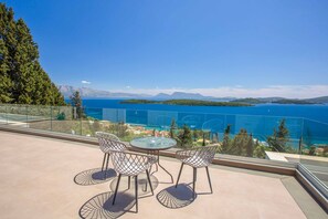 Un balcon doté d’une balustrade en verre, de deux chaises et d’une table, offrant une vue sur une étendue d’eau et sur des montagnes lointaines.