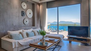 A modern living room with a large TV, a sofa, and a coffee table. There is a view of the ocean through large windows. - Villa Ino Lefkas - Four Bedroom Villa, Sleeps 8 (Perigiali)