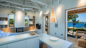 Une cuisine moderne avec des meubles blancs, un îlot central et une vue sur la mer par une porte ouverte.