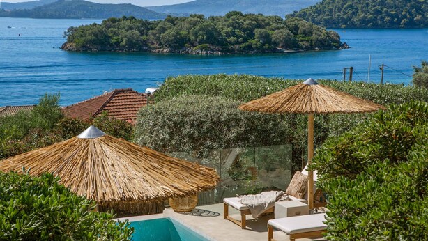Un espace piscine doté de chaises longues et d’un parasol en chaume, entouré de verdure et offrant une vue sur un lac avec une île au loin.