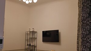 Un salon avec un lustre, un téléviseur fixé au mur, une table à manger avec des chaises et un canapé noir.