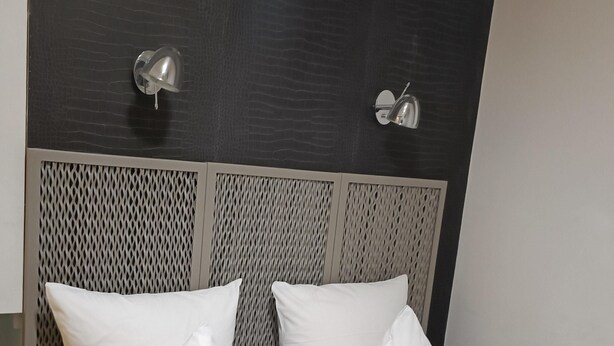 Un lit bien fait, avec des oreillers blancs et à rayures, une tête de lit à motifs texturés et deux lampes fixées au mur.