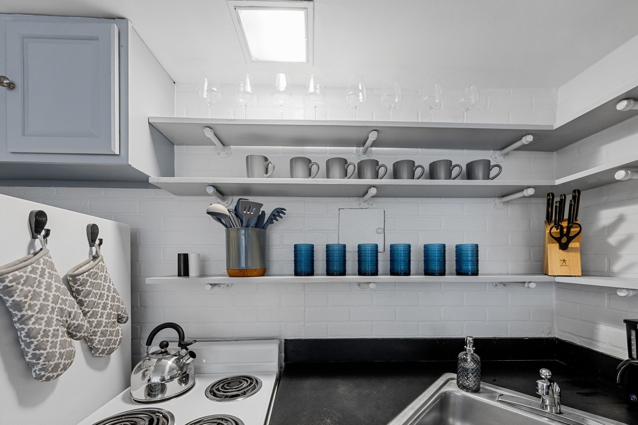 A kitchen with white brick walls, a stove, a sink, and shelves holding mugs and utensils.