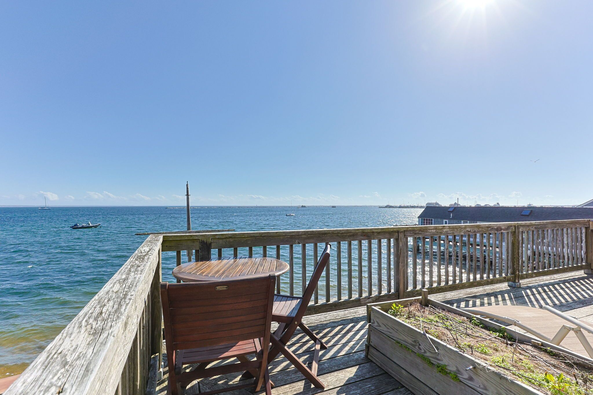 A wooden deck with a table and chairs overlooking a calm sea with boats.
