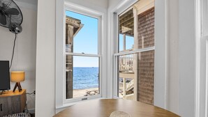 Une pièce avec une table en bois, un tapis roulé, une lampe et un téléviseur posé sur une commode en bois. De grandes fenêtres offrent une vue sur l’océan et donnent sur une terrasse en bois.