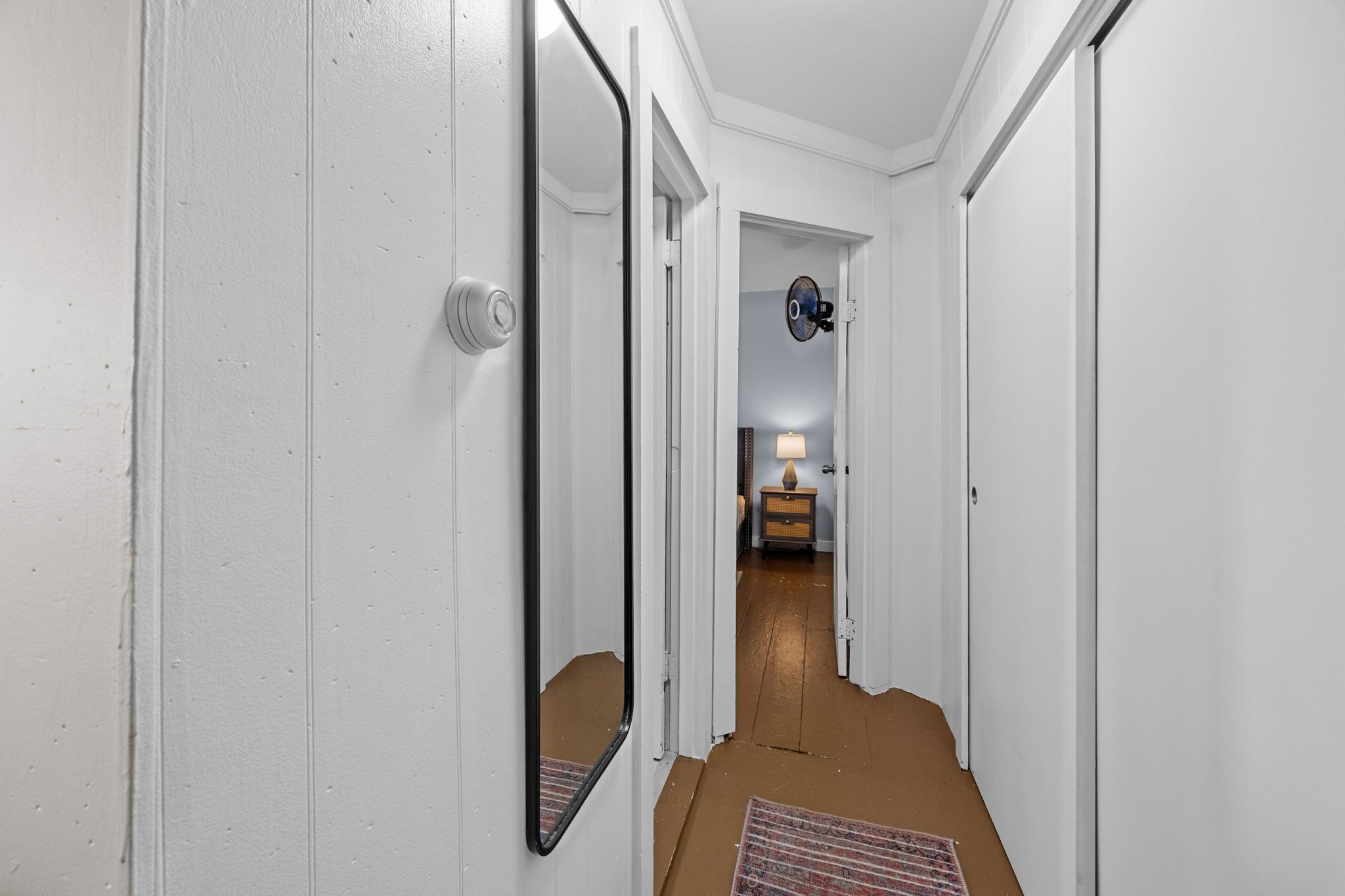 Un pasillo con paredes blancas y una puerta que da a una habitación con cama y lámpara.