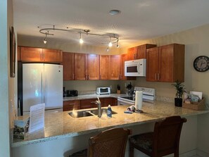 Una cocina con armarios de madera, un refrigerador de acero inoxidable, un microondas, un lavamanos y una encimera con diversos electrodomésticos y objetos.