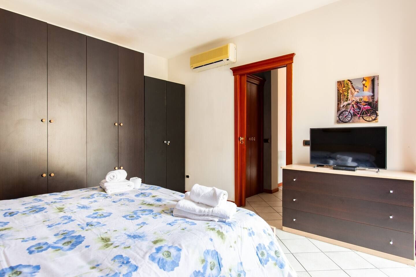 A bedroom with a bed, dark wooden wardrobe, a television on a dresser, and a painting on the wall.