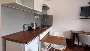 Une petite cuisine équipée comprenant un réfrigérateur, un évier et un plan de travail en bois. On y trouve une chaise blanche et une petite table à manger avec une chaise. Un téléviseur est fixé au mur.