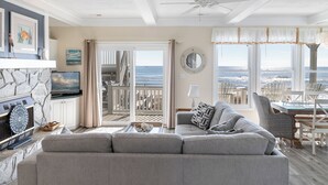 A living room with a large gray sofa, a television, a fireplace, and a view of the ocean through large windows. - Pier Pointe 3B2, Oceanfront Condo, in Emerald Isle, NC! (Emerald Isle)