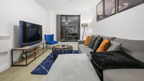 Ein modernes Wohnzimmer mit einem Flachbildfernseher, einem grauen Sofa mit orangefarbenen und schwarzen Kissen, einem blauen Sessel, einem runden Couchtisch und einem blauen Teppich.