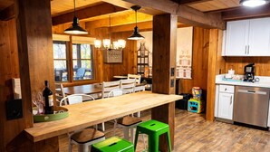Un intérieur en bois avec une longue table, des tabourets verts, des luminaires suspendus et un coin cuisine.