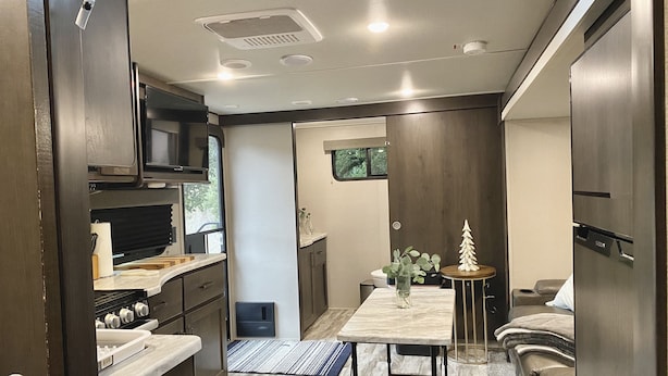 A compact kitchen with a microwave, stove, and sink. There's a dining table with a vase and a small tree decoration. The room has a wooden floor and a window with a view of greenery. - Relax to the Sound of the River while you soak in a Private Hot Tub  (Carmel Valley)