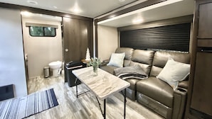 A compact RV interior with a kitchenette, a seating area with a sofa and cushions, and a small table. - Relax to the Sound of the River while you soak in a Private Hot Tub  (Carmel Valley)