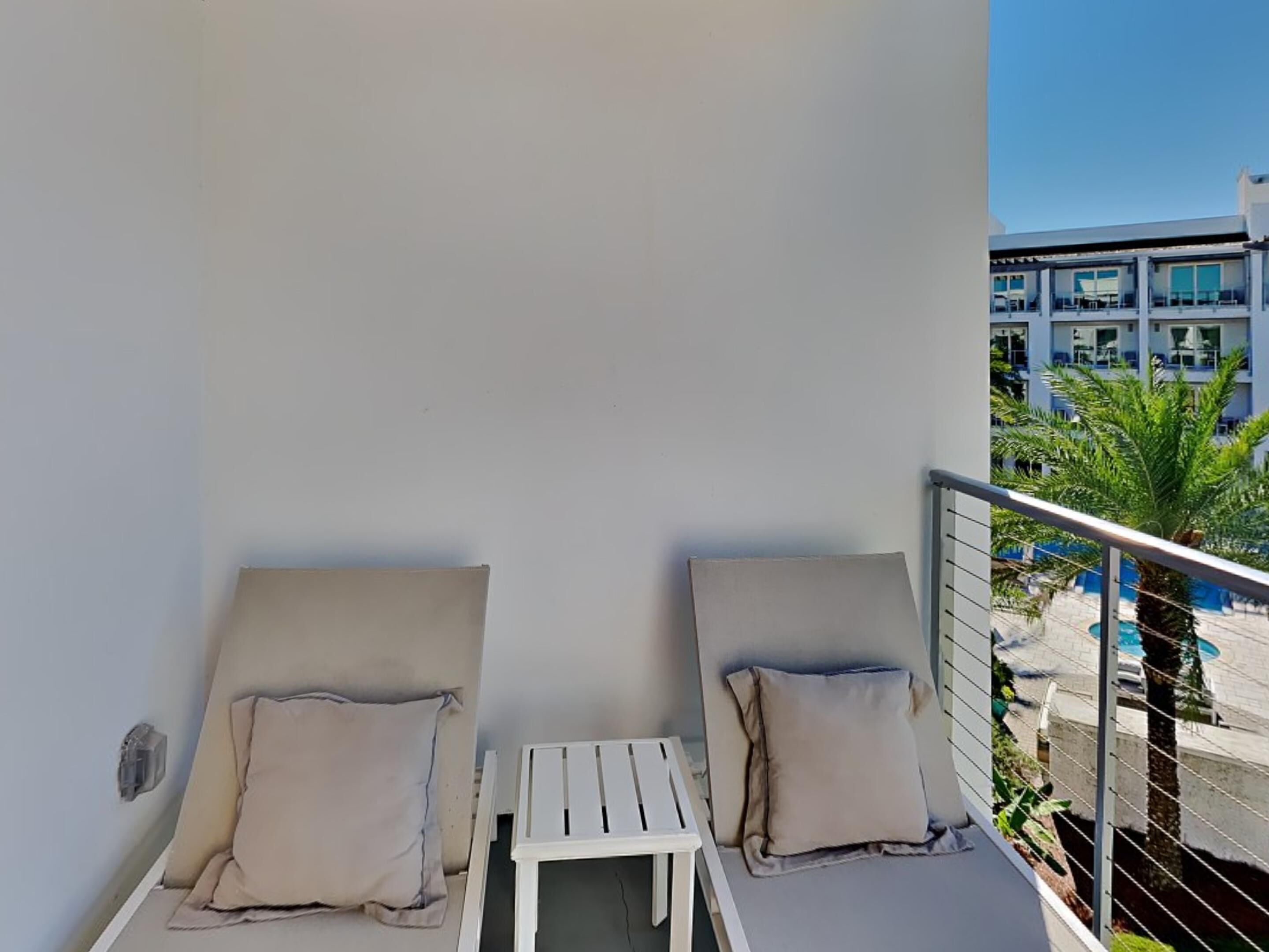 En balkong med två vita solstolar och ett litet bord, med utsikt över en pool och en byggnad med flera balkonger.