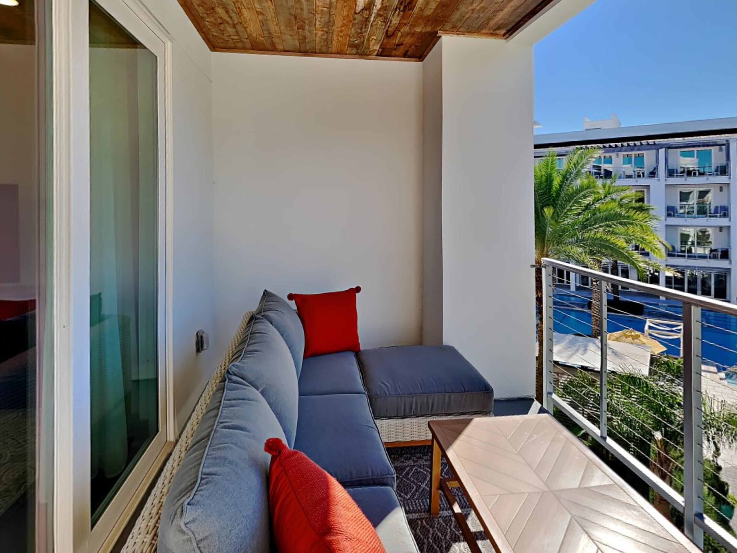 A balcony with a sofa, red cushions, and a view of a pool and buildings.