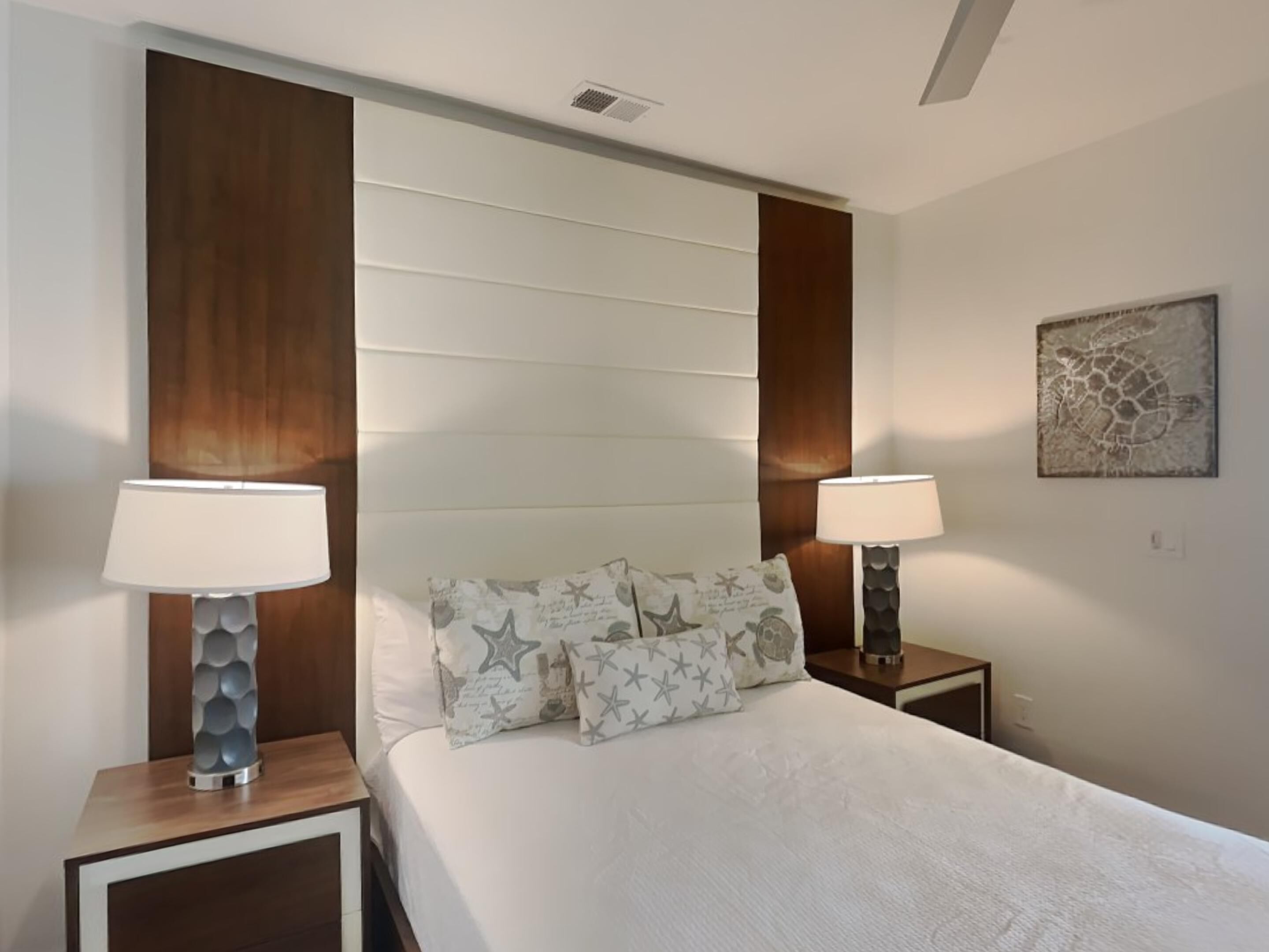 A bedroom with a large bed, two bedside tables with lamps, a headboard, and a wall-mounted artwork.
