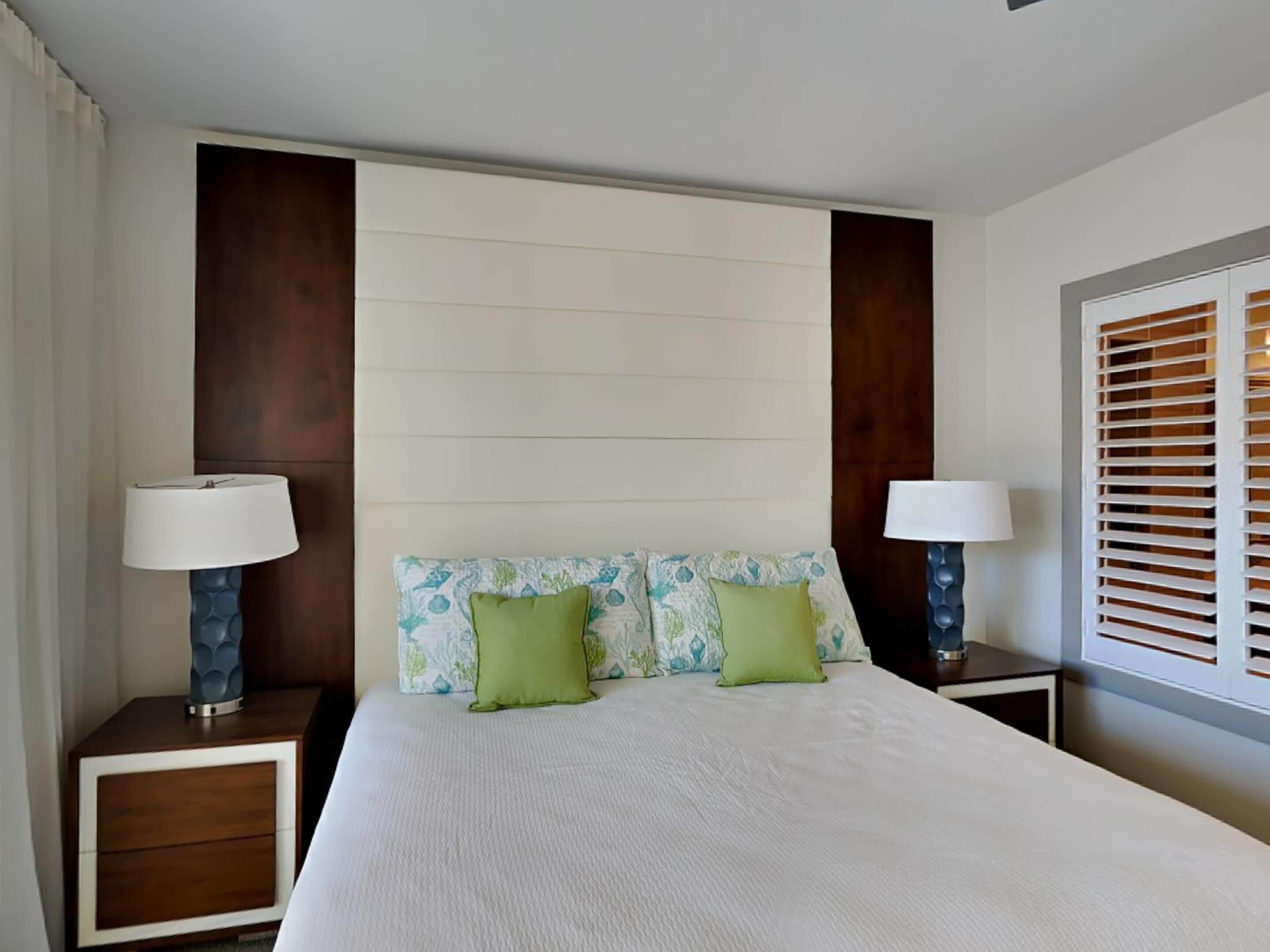 A hotel room with a large bed, two bedside tables with lamps, a window with blinds, and a headboard with a floral pattern.