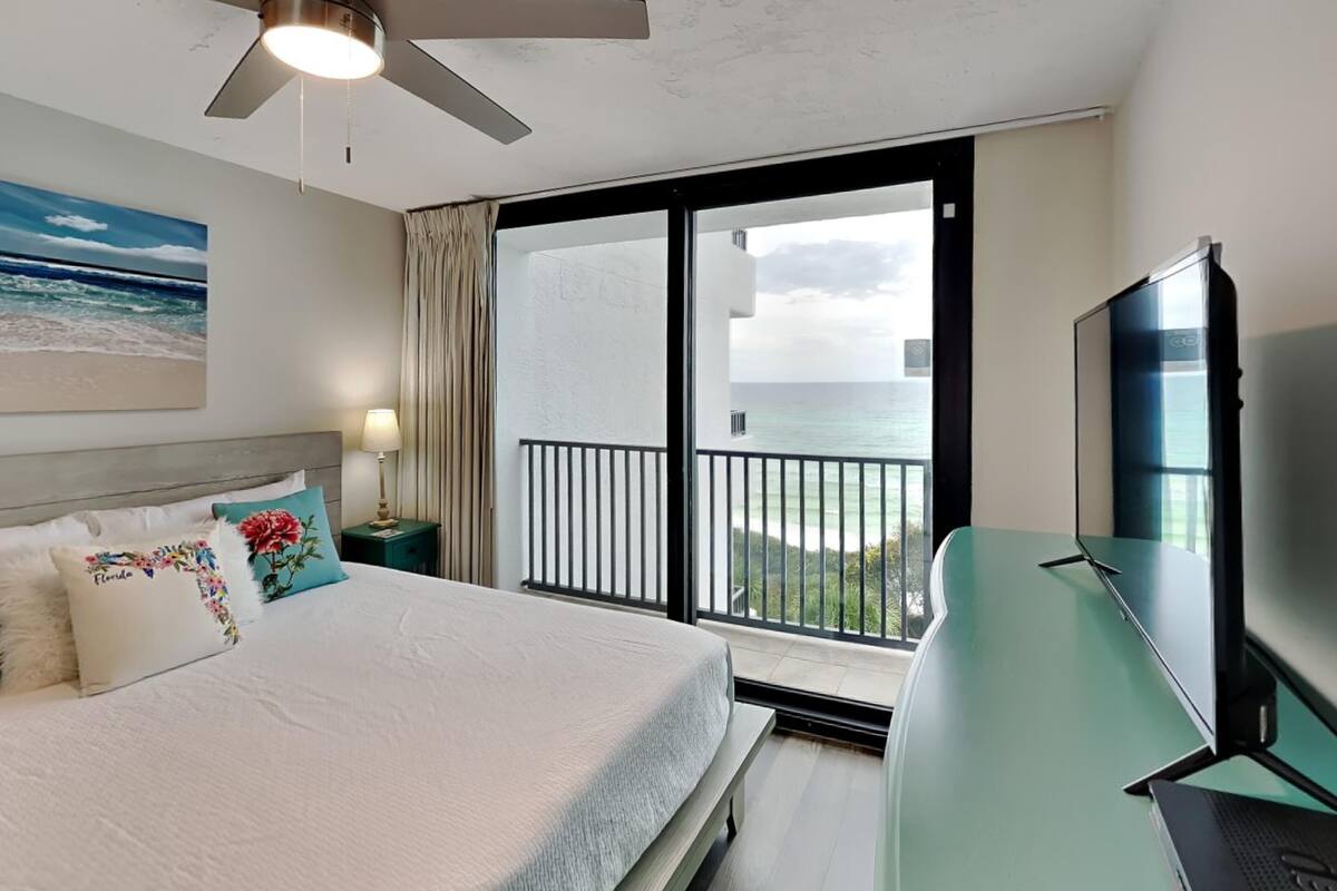 A hotel room with a bed, a TV, a ceiling fan, and a balcony with a view of the ocean.