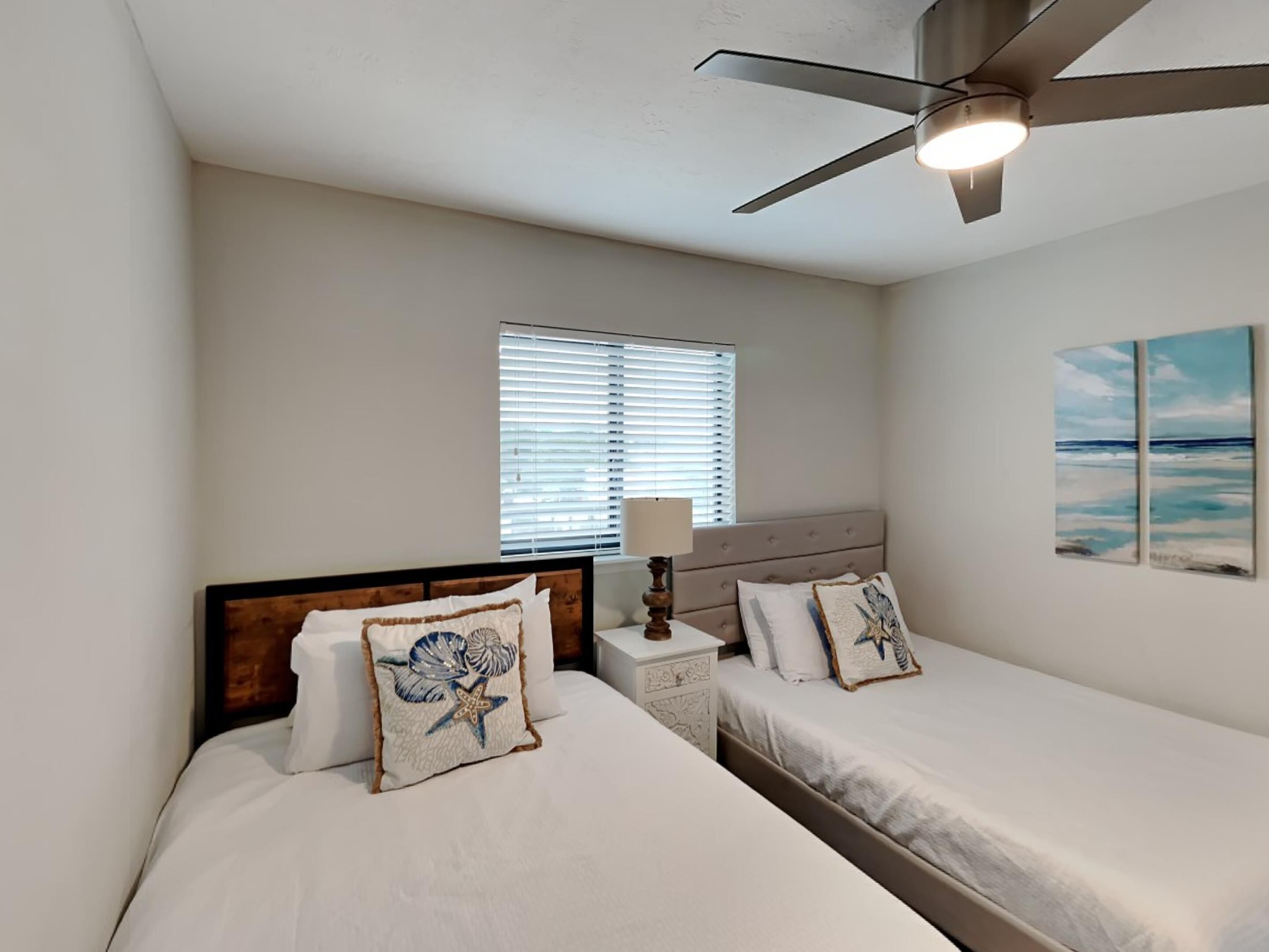 A bedroom with two beds, a ceiling fan, a window with blinds, and a painting on the wall.