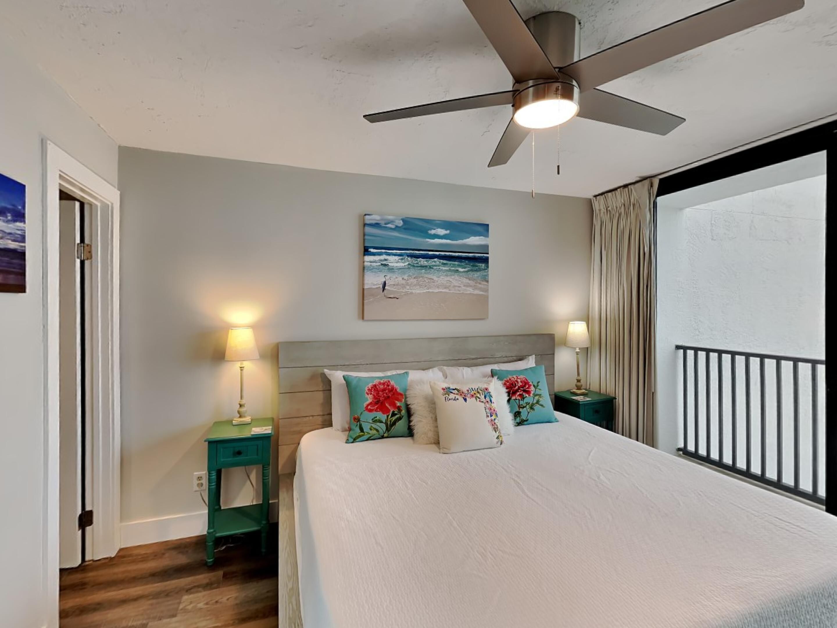 A bedroom with a bed, a ceiling fan, a nightstand with a lamp, a painting of a beach scene, and a view of the ocean through a window.