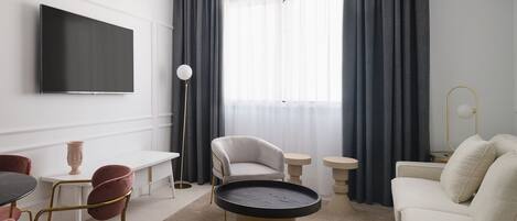 Ett modernt hotellrum med en platt-TV, ett runt soffbord, en soffa, ett matbord med stolar och en golvlampa.