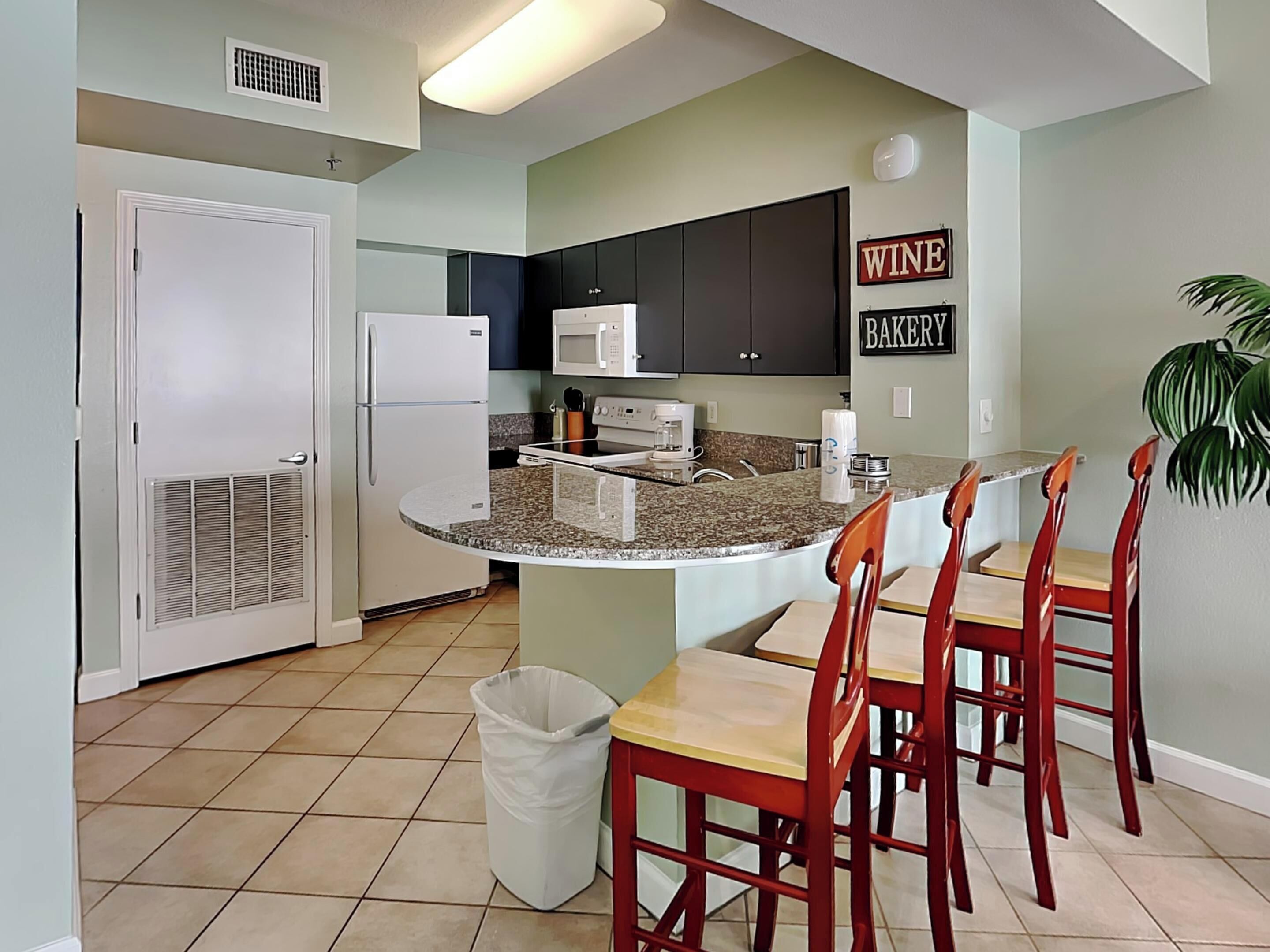 A kitchen with a central island, a microwave, a refrigerator, and a trash can. There are chairs around the island and a sign on the wall that says "WINE BAKERY".