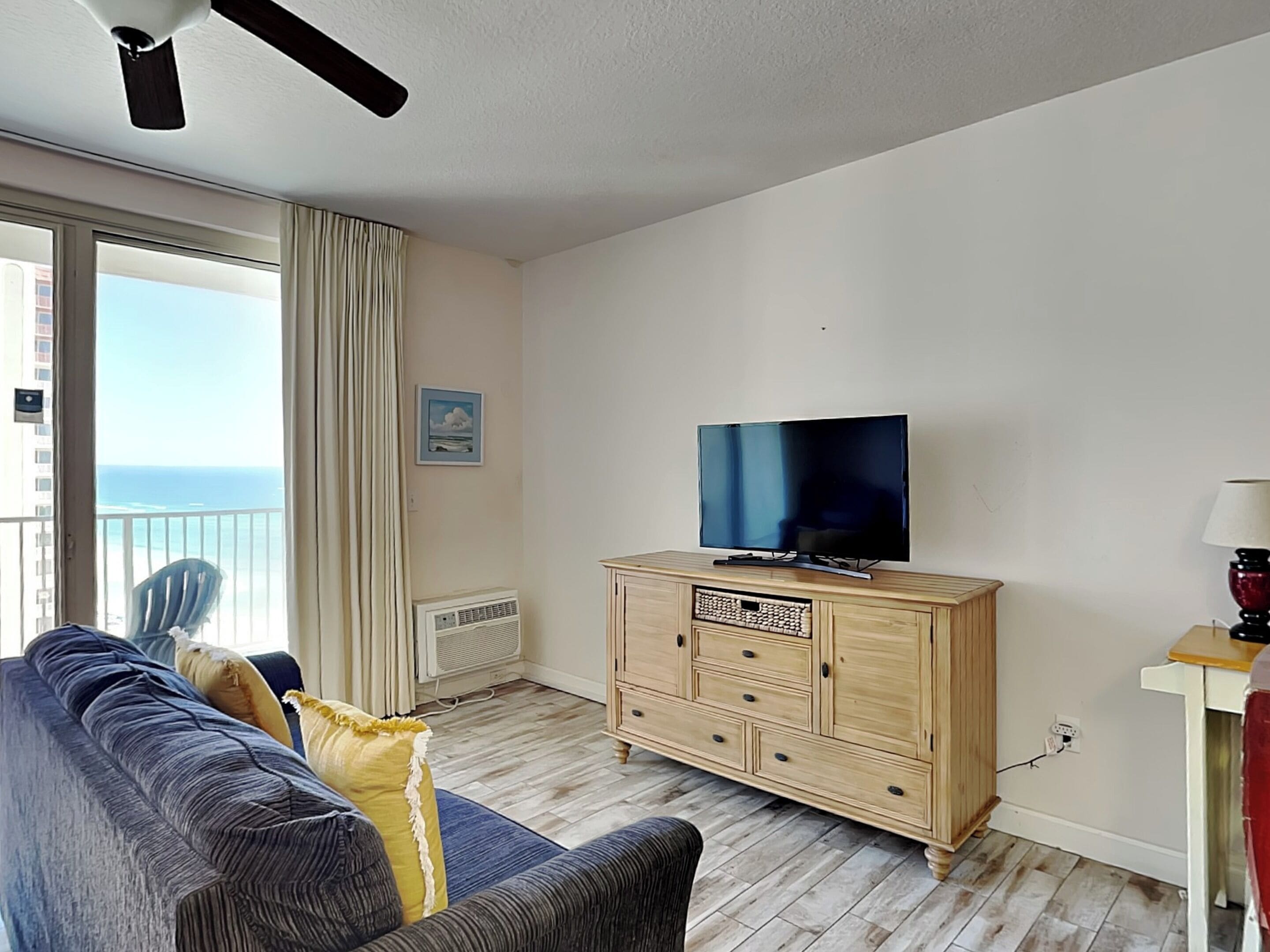 Una sala de estar con un televisor sobre un mueble de madera, un sofá, un sillón y vista al océano desde un balcón.