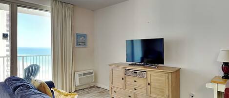 Una sala de estar con un televisor sobre un mueble de madera, un sofá, un sillón y vista al océano desde un balcón.
