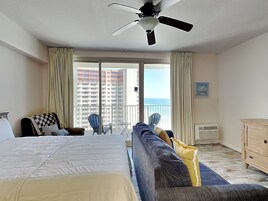 Ein Hotelzimmer mit Bett, Sofa, Balkon mit Blick auf Hochhäuser und das Meer sowie einem Deckenventilator.
