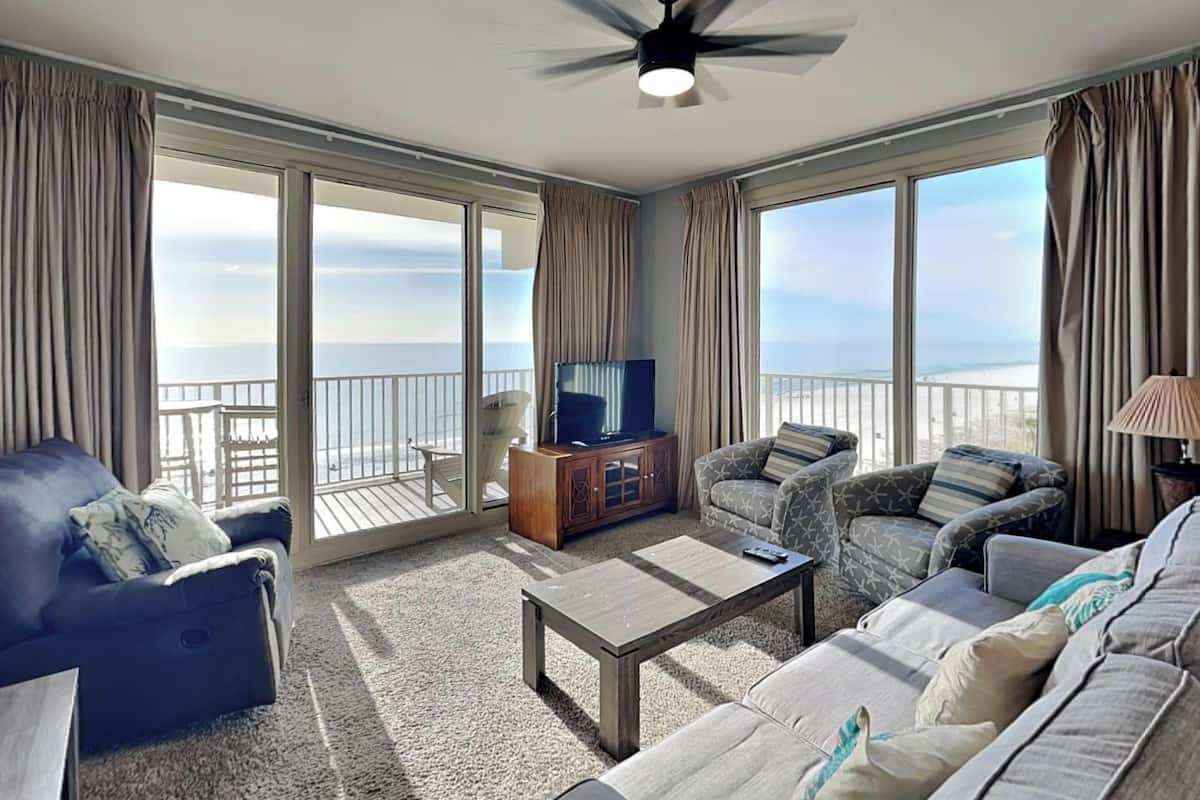 A spacious living room with a large sofa, a coffee table, and a TV on a wooden cabinet. The room has large windows with curtains, offering a view of the beach.