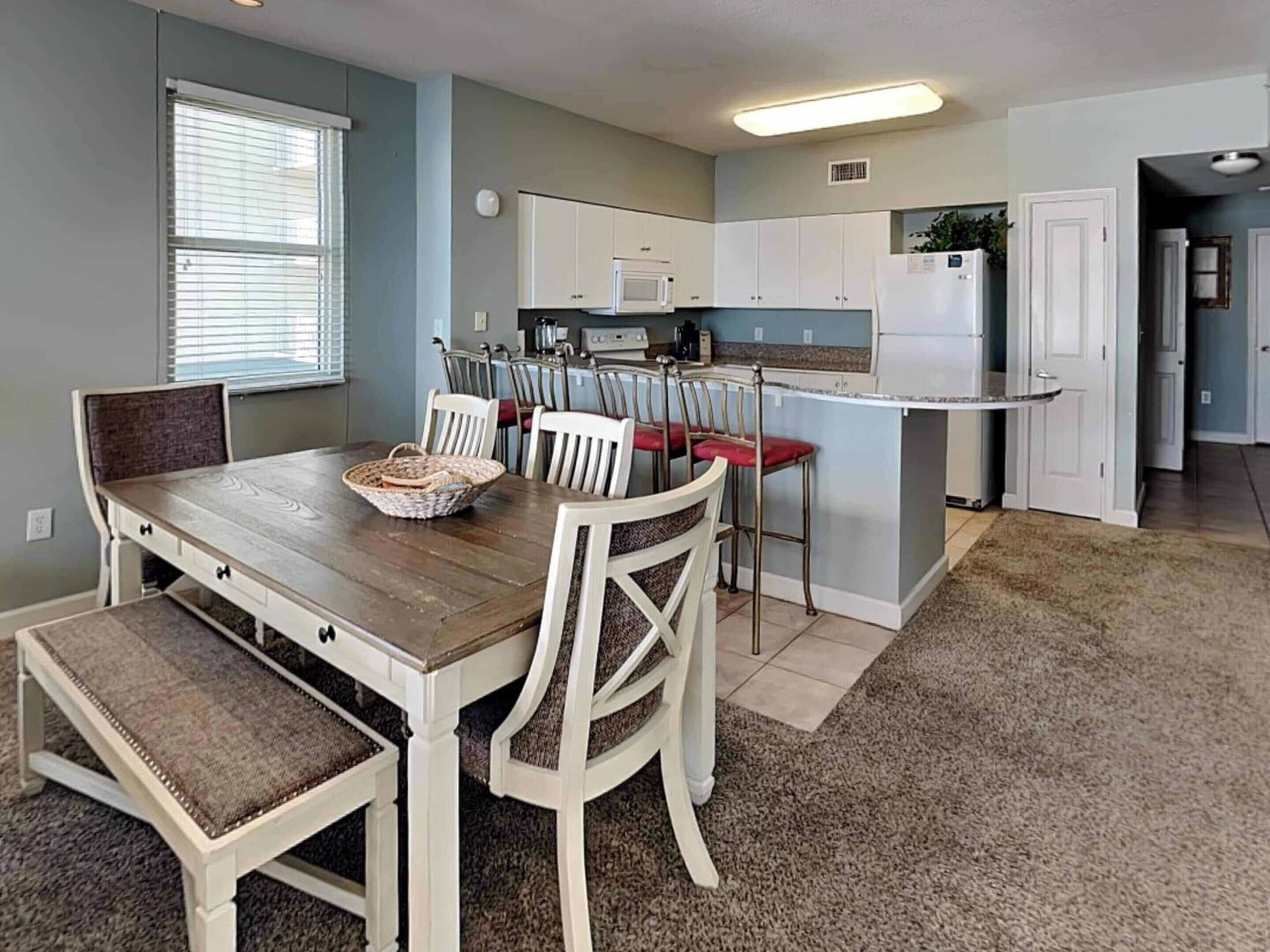 Un comedor con mesa y sillas de madera, una cocina con armarios blancos, microondas y refrigerador.