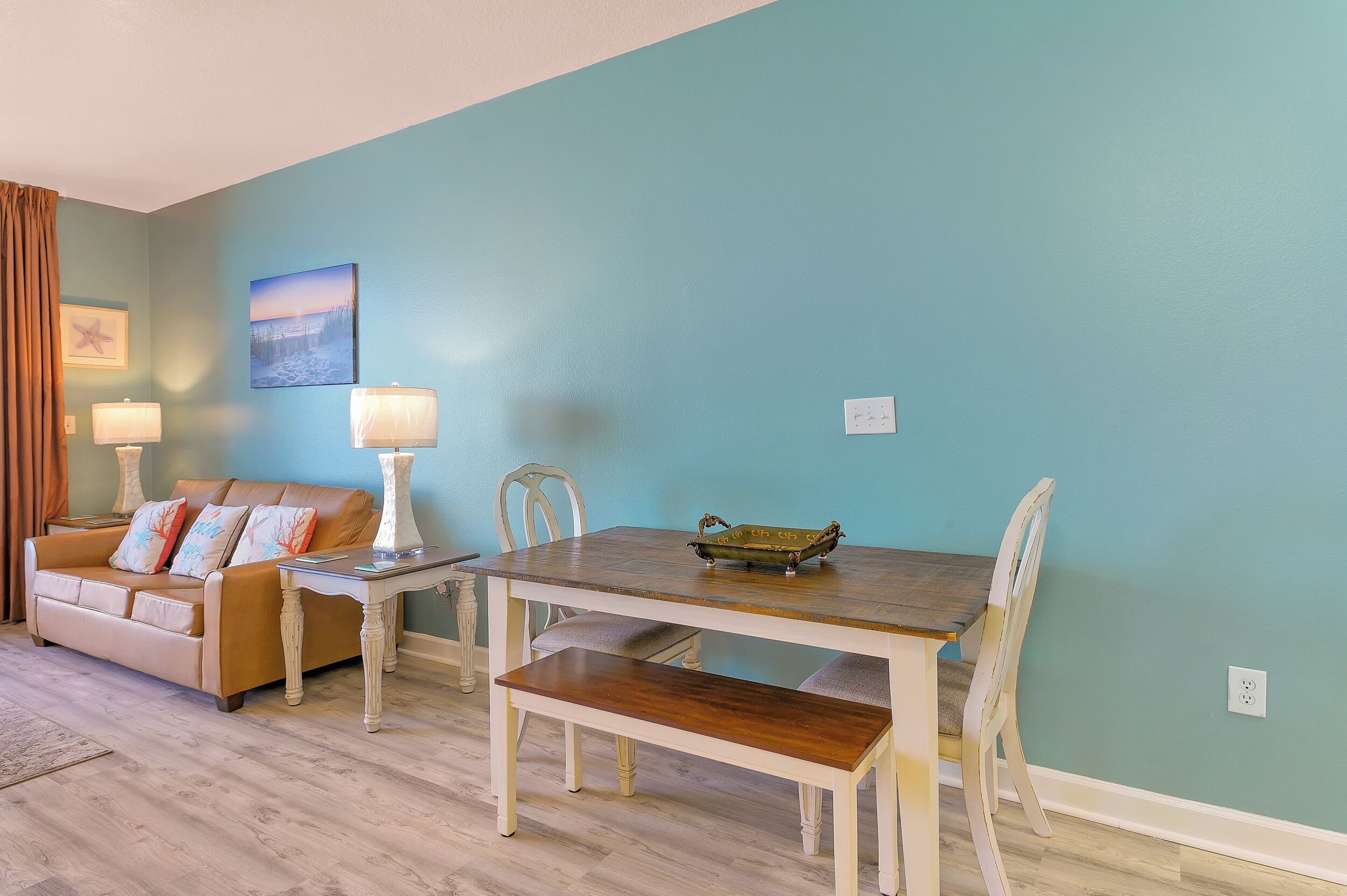 Uma sala de estar com parede azul, sofá castanho, mesa de jantar de madeira com cadeiras e duas luminárias.